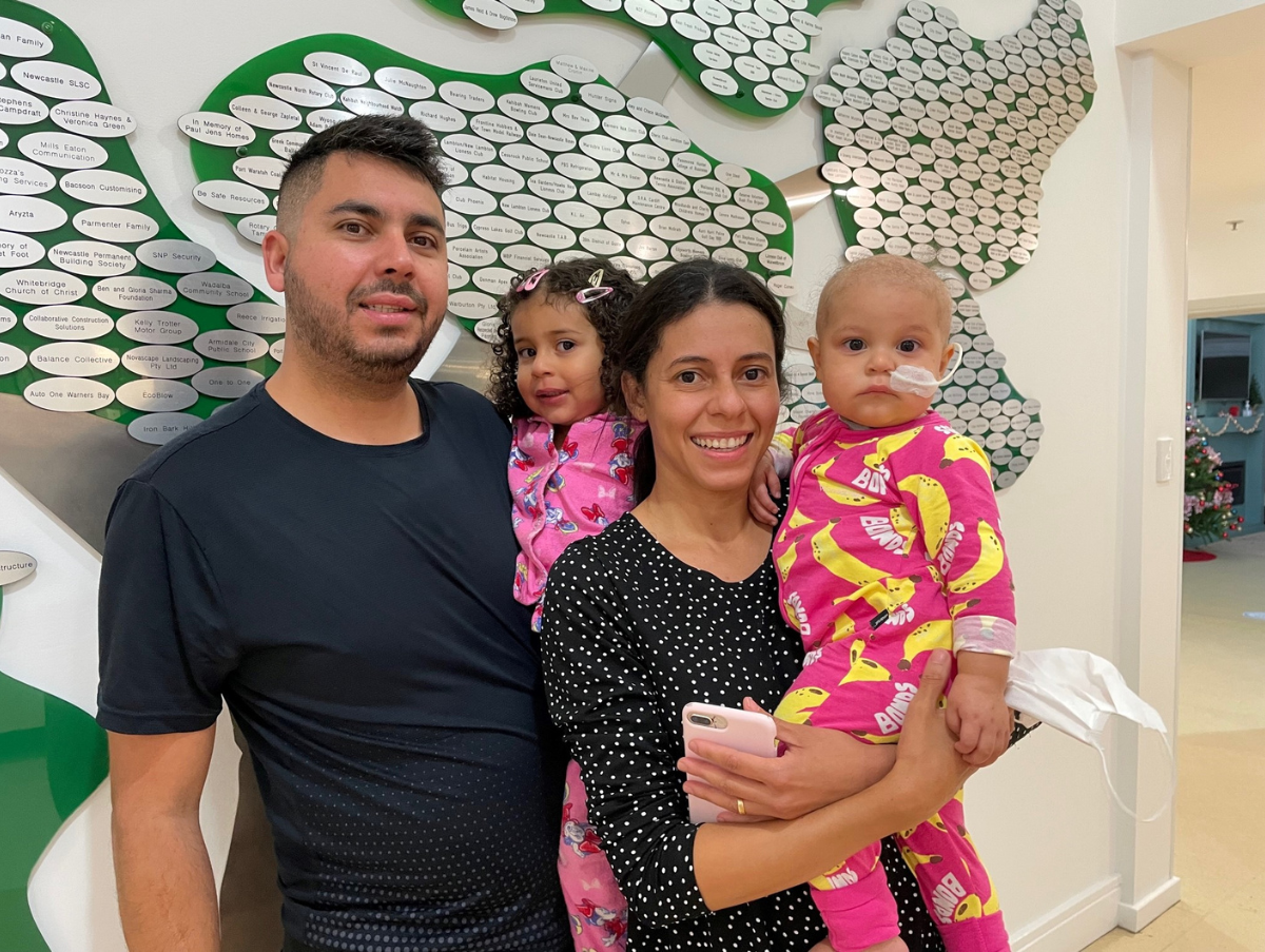 A family standing in front of the Tree of Love smiling at the camera. The dad is holding his small daughter. And the mum is holding another small daughter who is wearing a nasal tube.