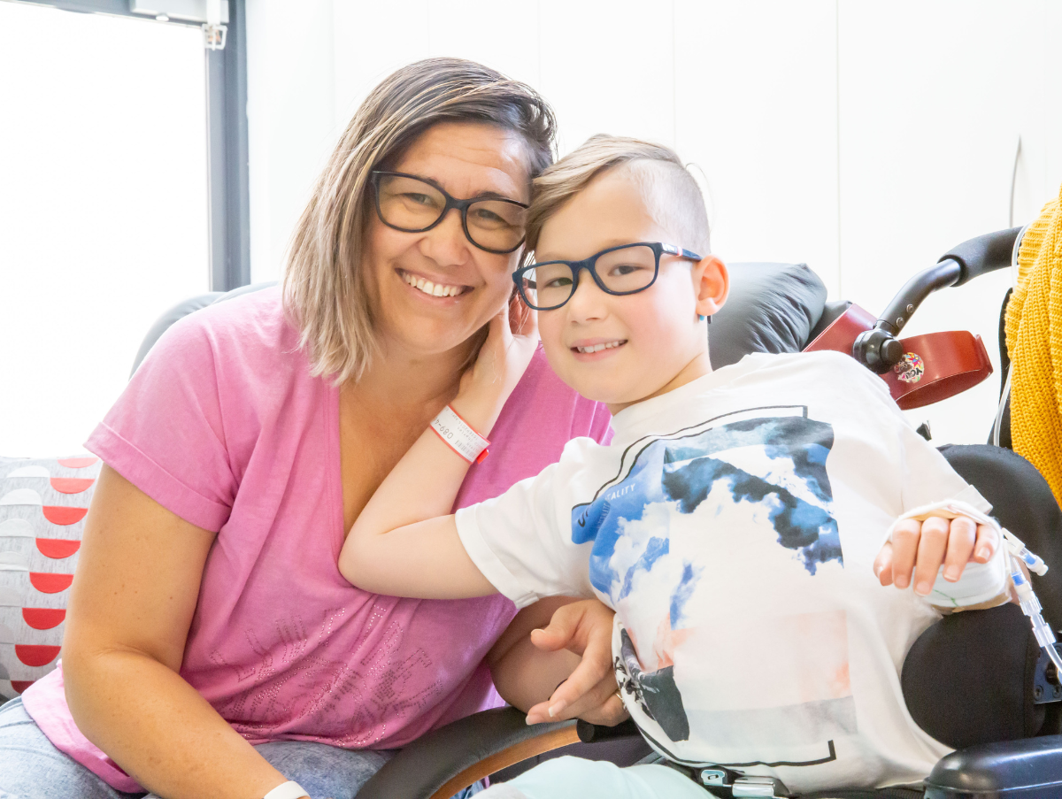 A mother and her son are smiling at the camera. The mother is wearing a pink shirt, black framed glasses and has blonde hair. The son also has black framed glasses and is sitting in a wheel chair. He has a hospital band on his right hand and his left hand is in a cast.