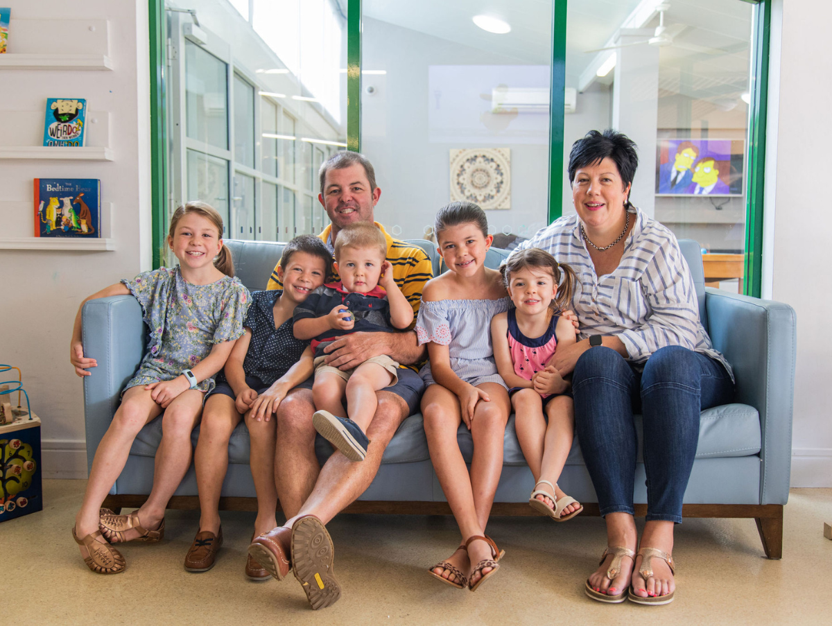 A family sitting on a blue couch at Ronald McDonald House Newcastle. The family (A mum, dad and five kids, including three girls and two boys all of various ages) are all smiling at the camera.