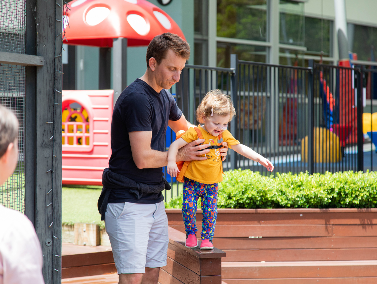 A dad helping his toddler daughter balance on a retaining wall in the playground at Ronald McDonald House Newcastle. The daughter has a nasal tube.