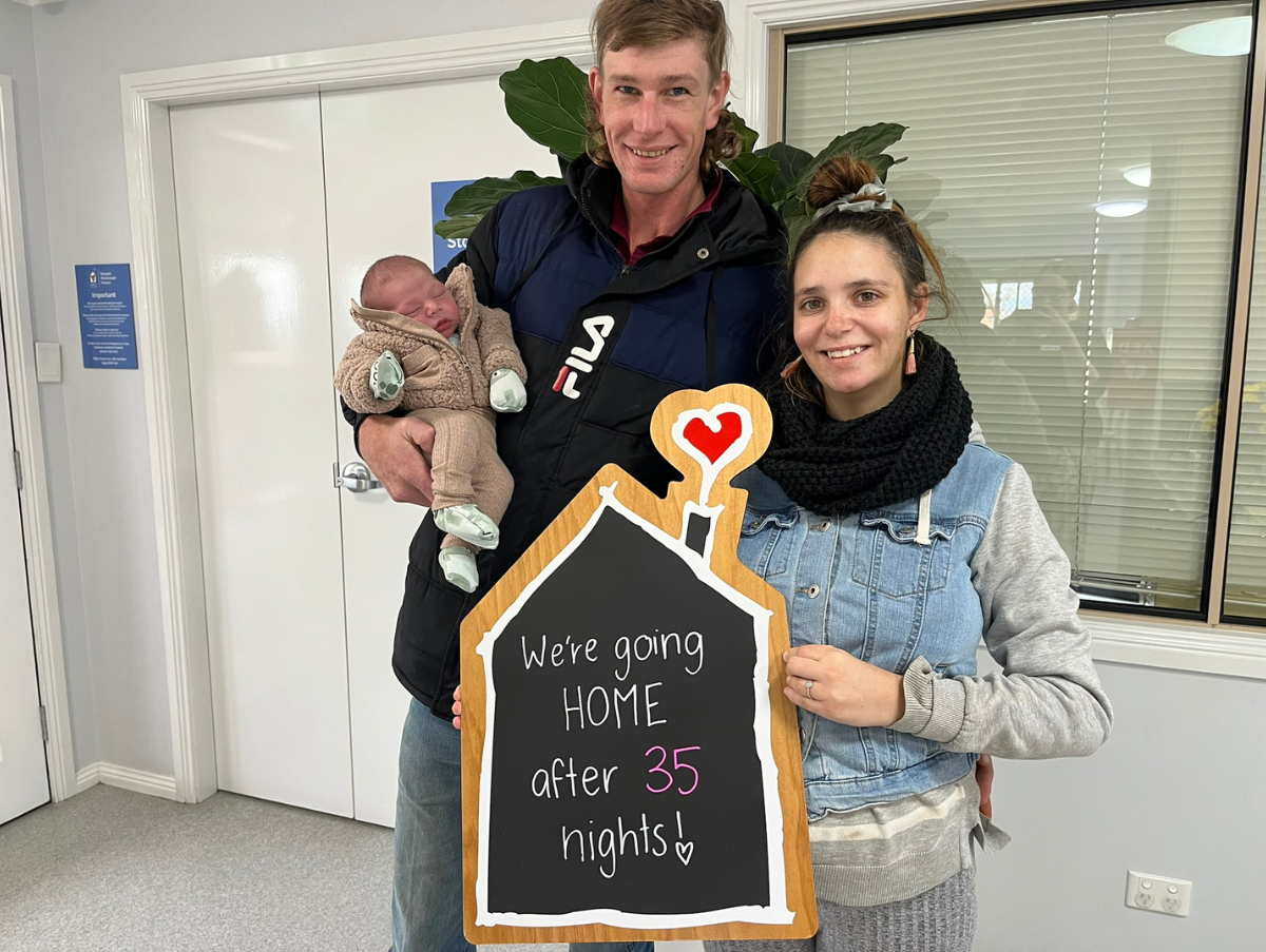 A mum and dad are standing in Ronald McDonald House Tamworth. The dad is holding a new born baby. And the mum is holding a sign which reads "We're going HOME after 35 nights!"