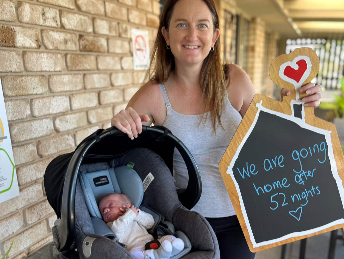 A mum is smiling at the camera holding a baby carrier with her new born baby inside. In her other hand, the mum is holding a chalk board sign which reads "We are going home after 52 nights"