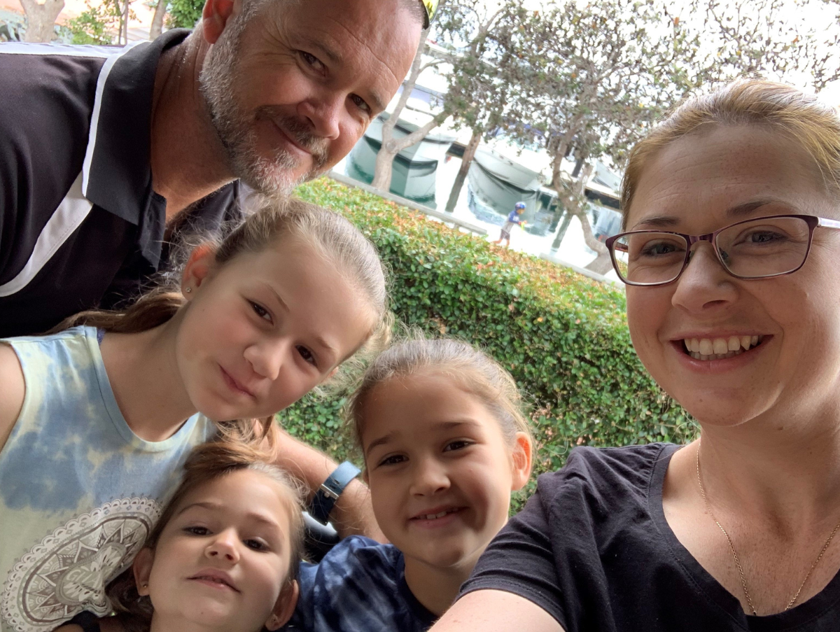 A mum (white, light brown hair and wearing glasses) is taking a selfie. In the photo are three daughters and their dad.