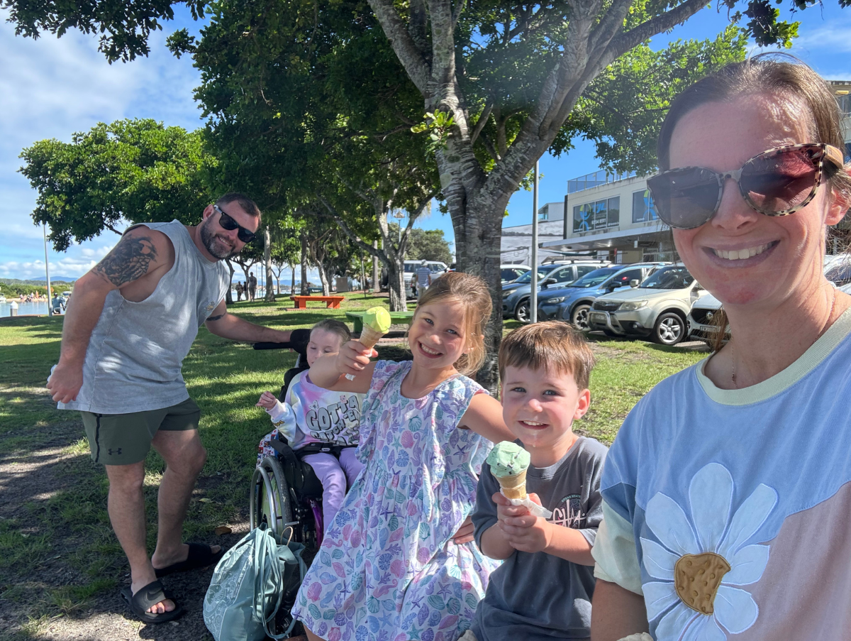 A mum and dad and their three children are sat on a grassy patch. Two of the children are eating icecream in a cone. One child is in a wheelchair. All are smiling at the camera.