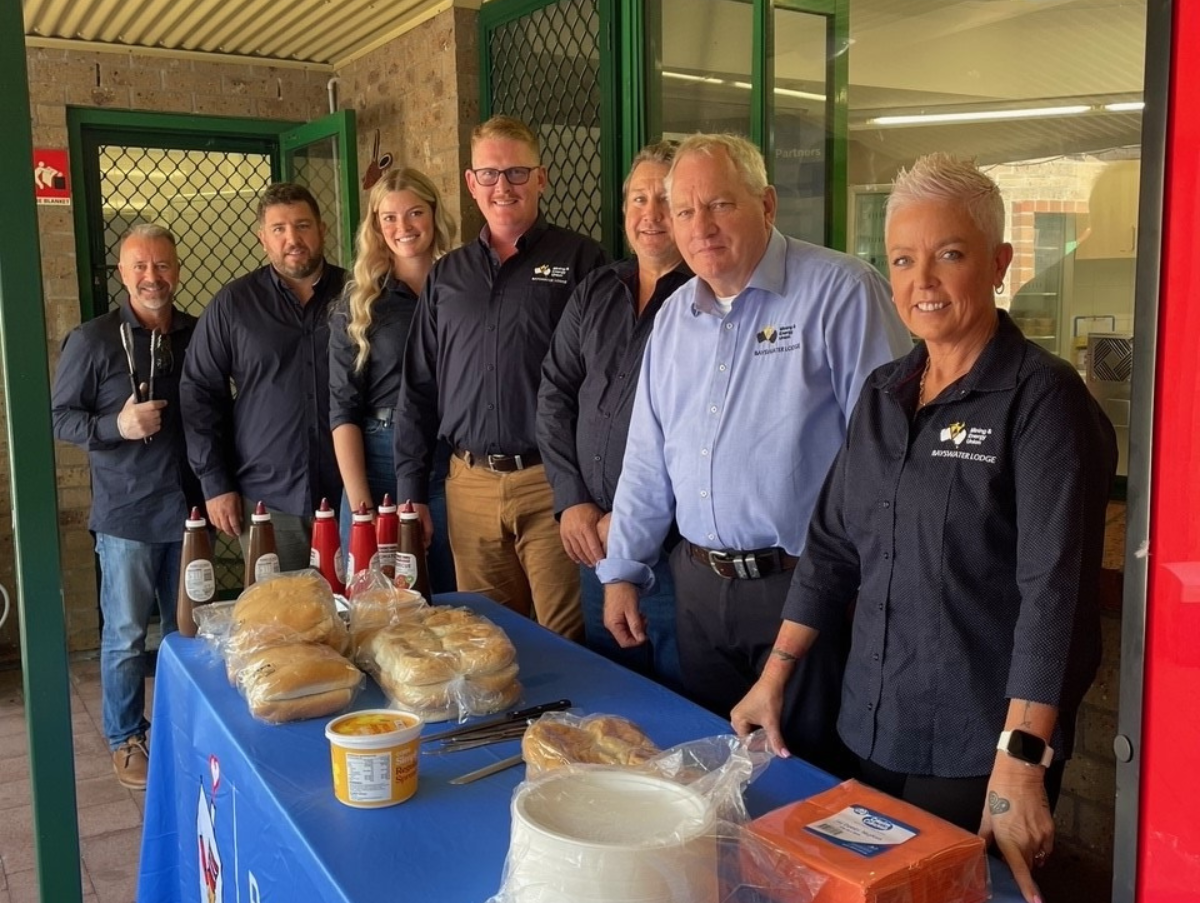 Seven people standing in front of a blue table. On the table are sauce bottles, bread rolls, butter, knives, plates and napkins. All seven people are smiling at the camera.