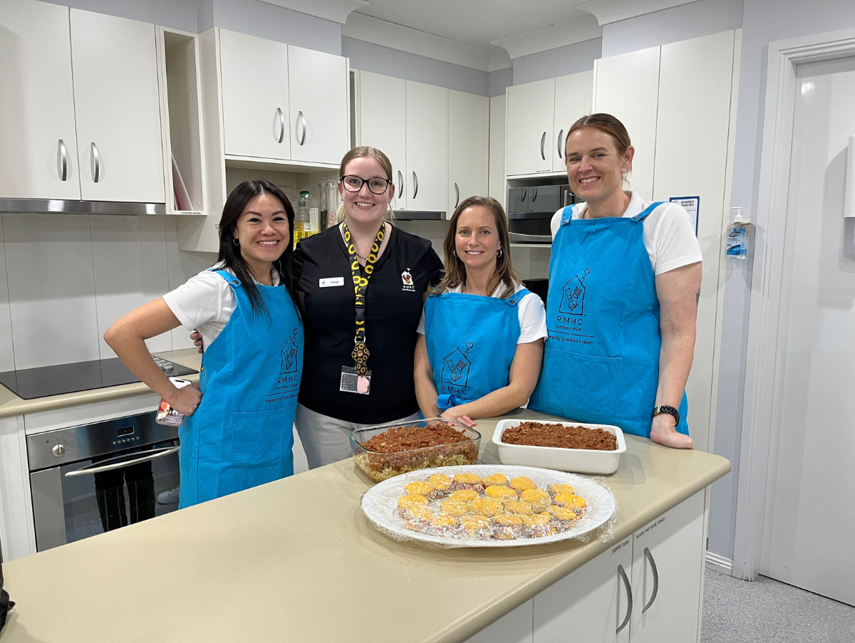 Four ladies standing in a white kitchen. Three are wearing blue cooking aprons. On a bench in front of them is a plate of melting moments and two baking dishes filled with lasagne bake.