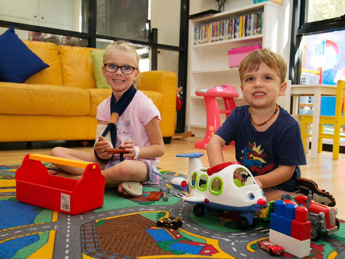 Two kids is sitting in the Ronald McDonald J1 Family Room. Both are playing with a toy plane and tractor. The young girl is wearing black framed glasses and has her right arm in a sling and cast.