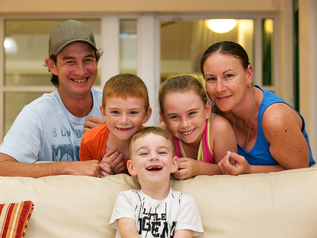 A family (mum, dad, two sons and a daughter) are leaning over a lounge smiling at the camera.