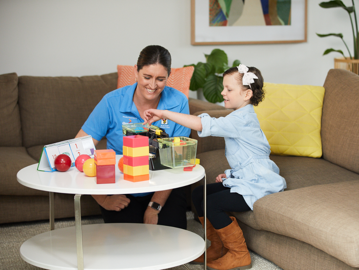 A Ronald McDonald Learning Program team member sitting on a lounge with a young child. The pair are playing with bricks and balls. and both are smiling and looking happy