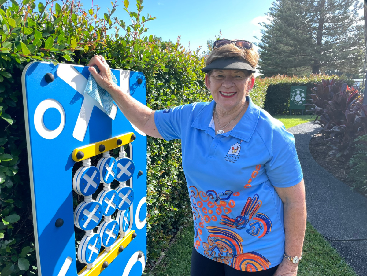 An older, white lady wearing a blue shirt and black cap with brown glasses ontop of her head smiles at the camera. The lady has on arm resting on a tall naughts and crosses board. Behind her is greenery and a footpath.