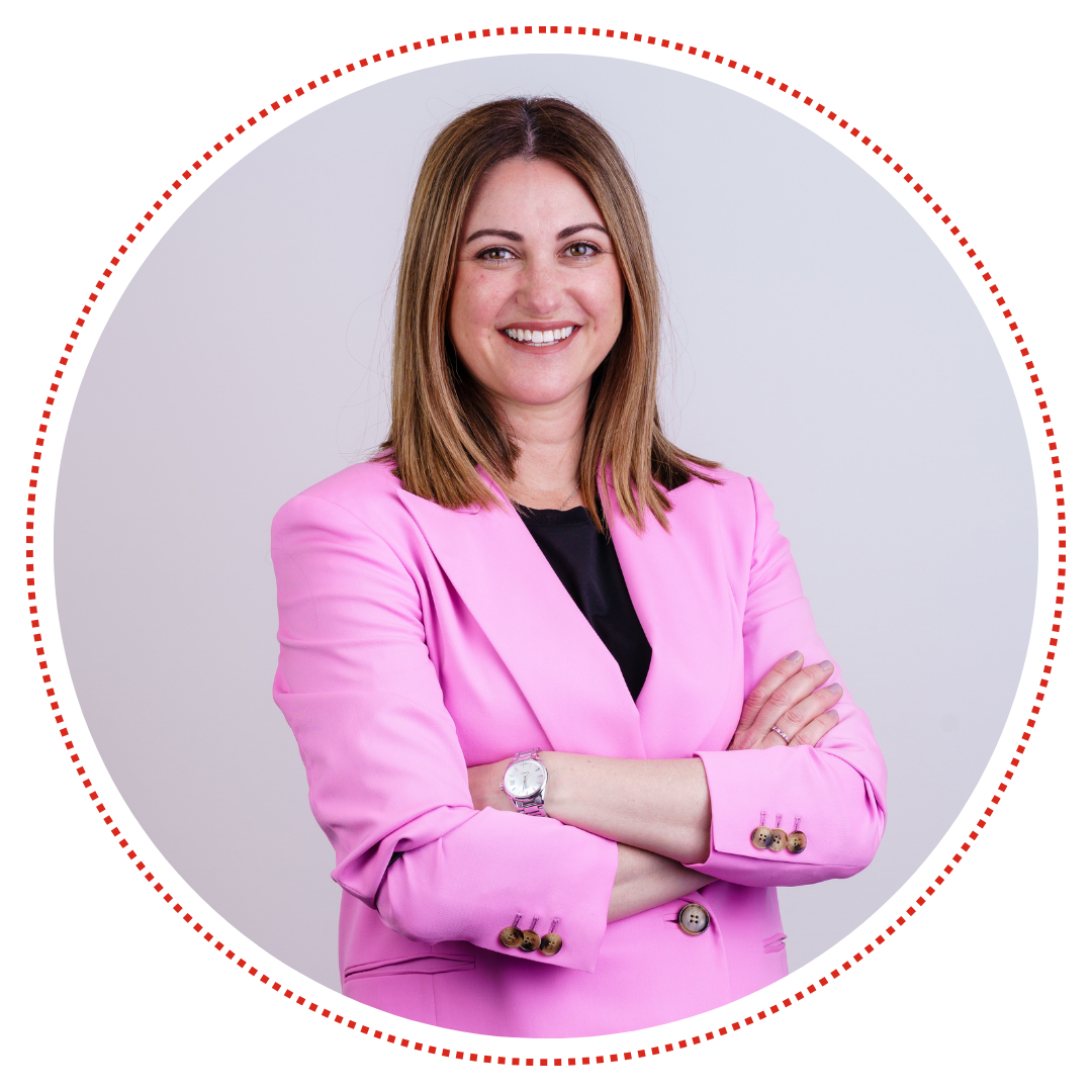 Mel, a white lady with brown hair, wearing a black shirt under a bright pink suit jacket, smiling at the camera