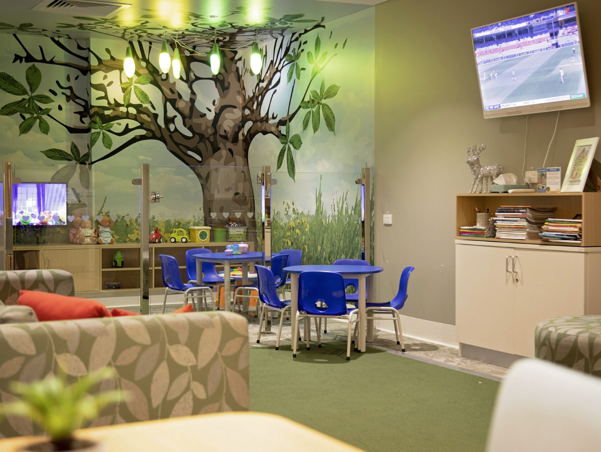 Wide room with a mural of a tree painted on the back wall. The carpet is green and in the room are kids tables and chairs, a green lounge, bookcase with books and a television.