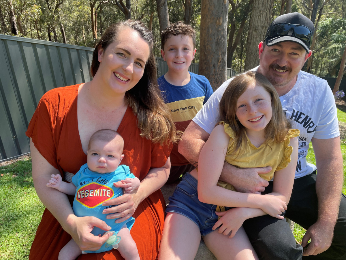 A family sitting on a stone bench outside. The mum is holding a new born baby, while the dad is cuddling a young girl. Behind them is a young boy. All are smiling at the camera