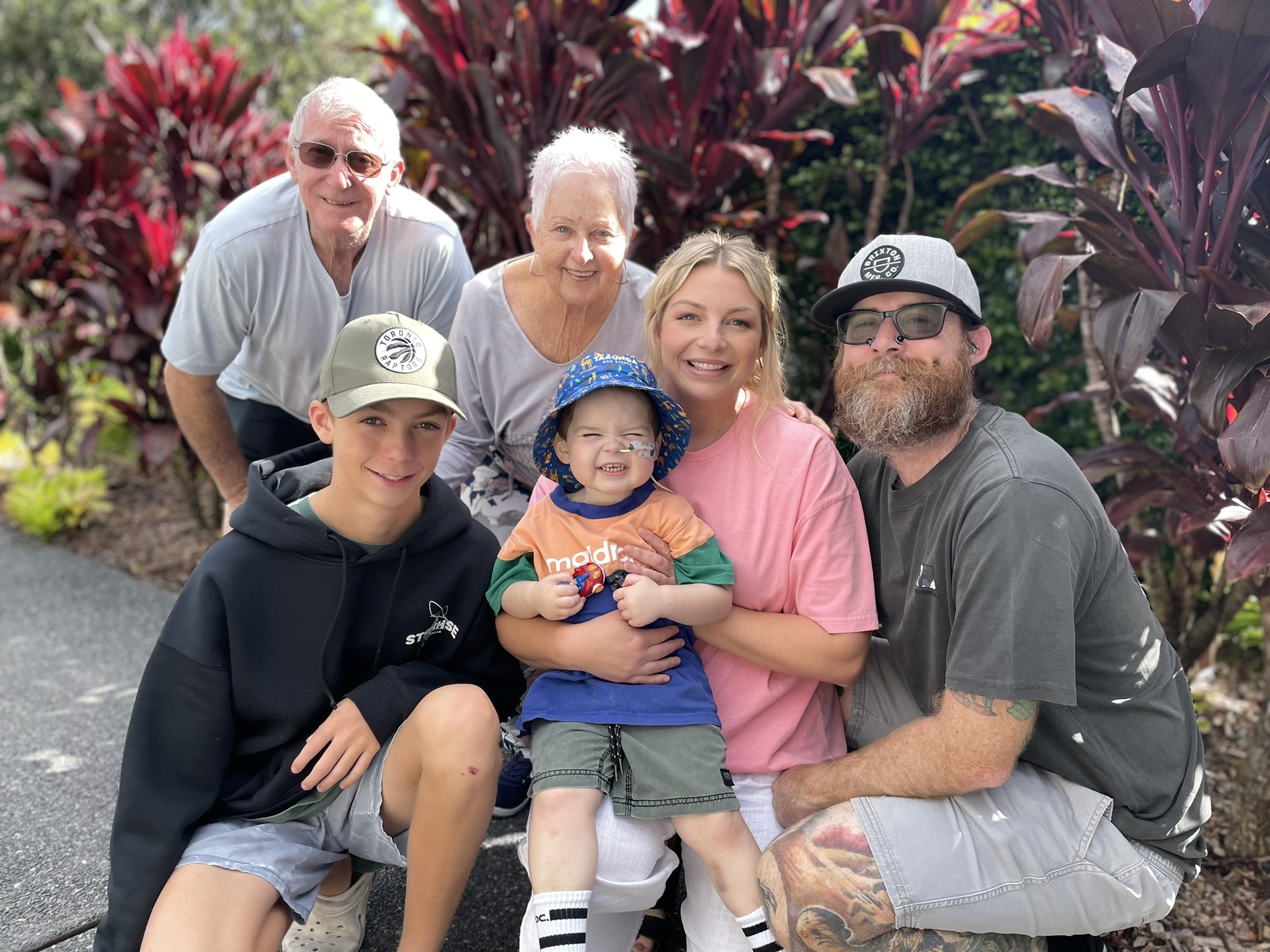 A family is crouched in front of greenery smiling at the camera. Included in the photo is a grandpa and grandma. In front of them is young boy, mum and dad. On mums lap is a small toddler wearing a nasal tube.