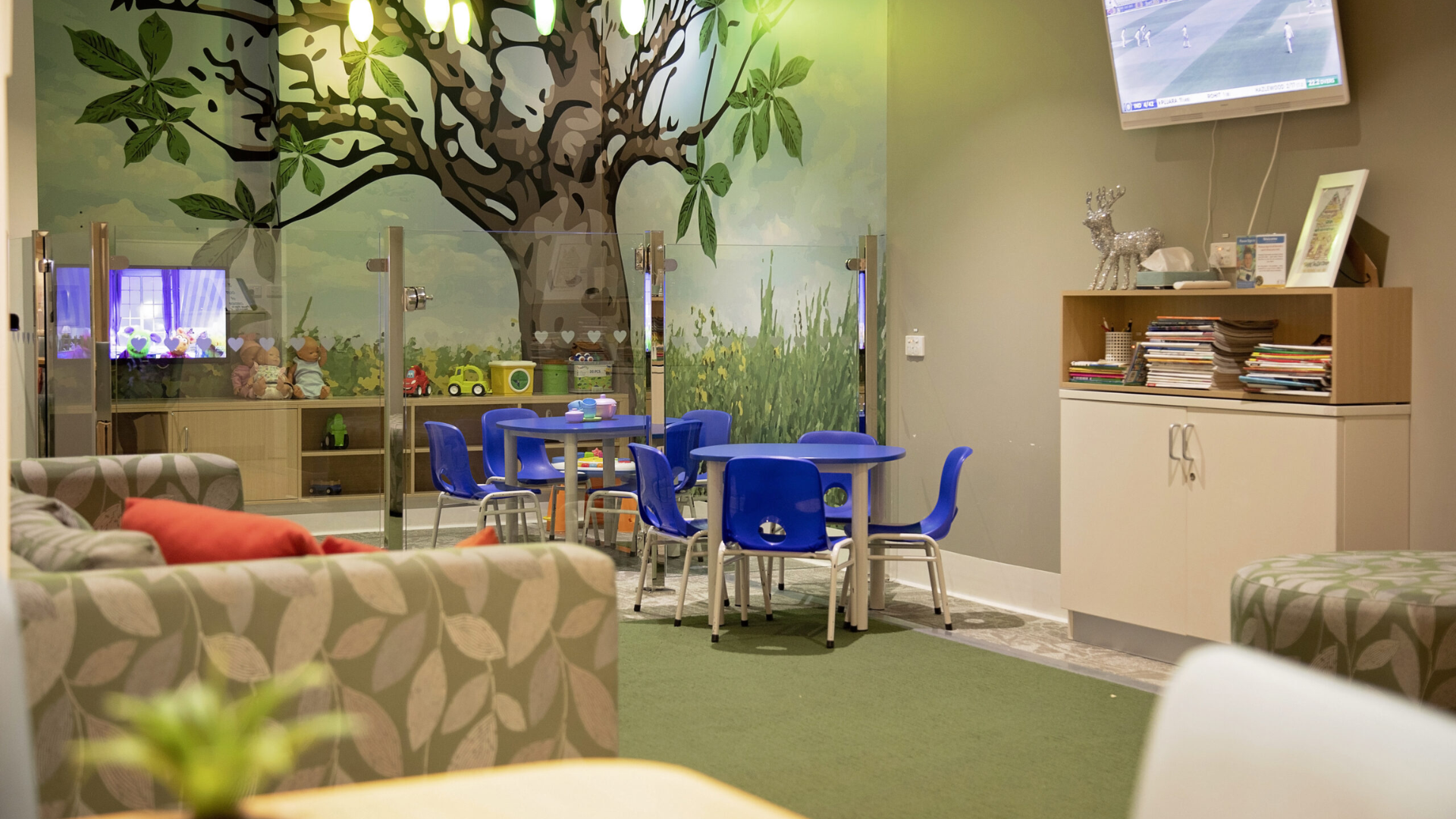 Wide room with a mural of a tree painted on the back wall. The carpet is green and in the room are kids tables and chairs, a green lounge, bookcase with books and a television.