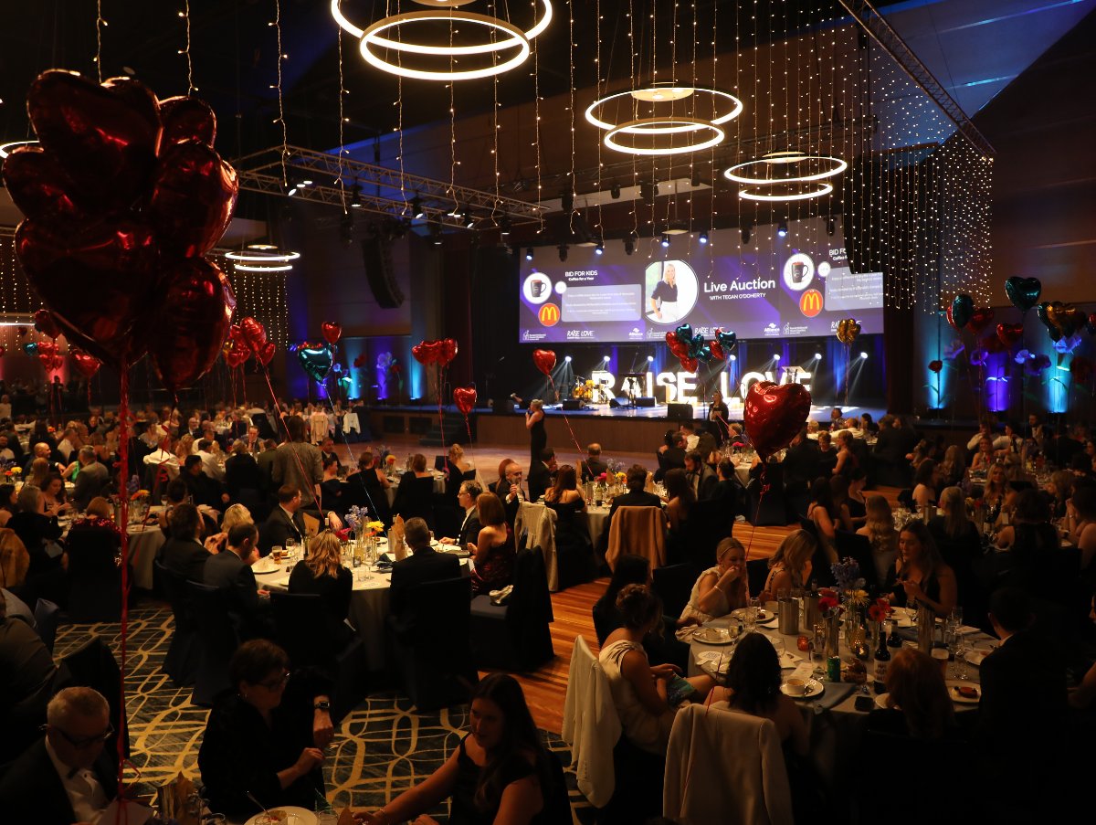 A function space at NEX Newcastle. The room has fairy lights handing from the ceiling, love heart balloons on tables around the room. People are sitting at tables watching the auctioneer infront of the stage