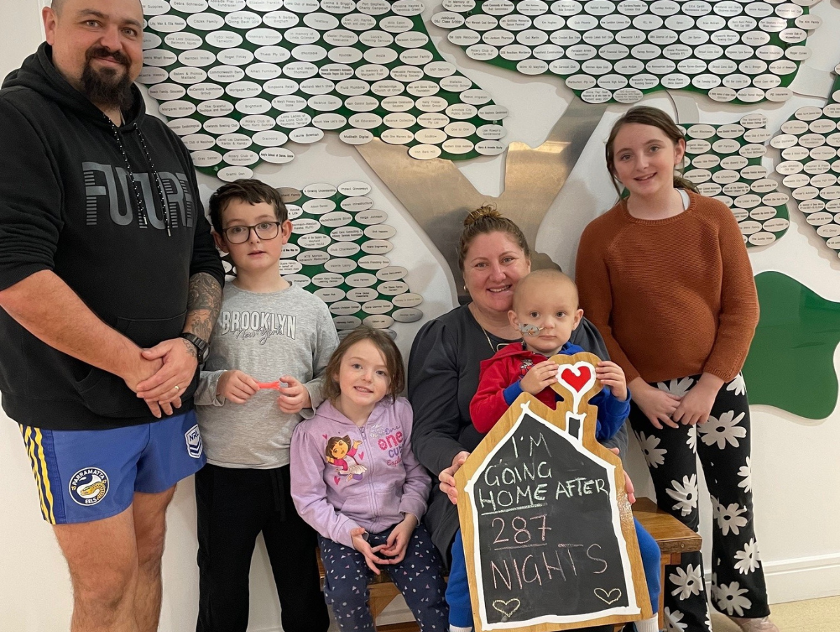 A family of six (mum, dad and four children) are standing together. All are smiling at the camera. The mum is holding a chalkboard sign which reads "I'm going home after 287 nights"