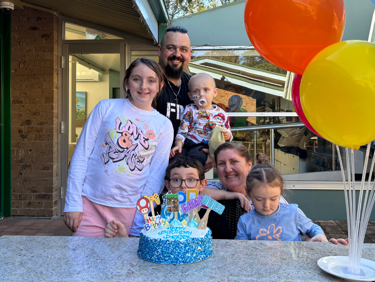 A mum and dad standing with their four children. On a table in front of them is a blue birthday cake and several bright coloured balloons.