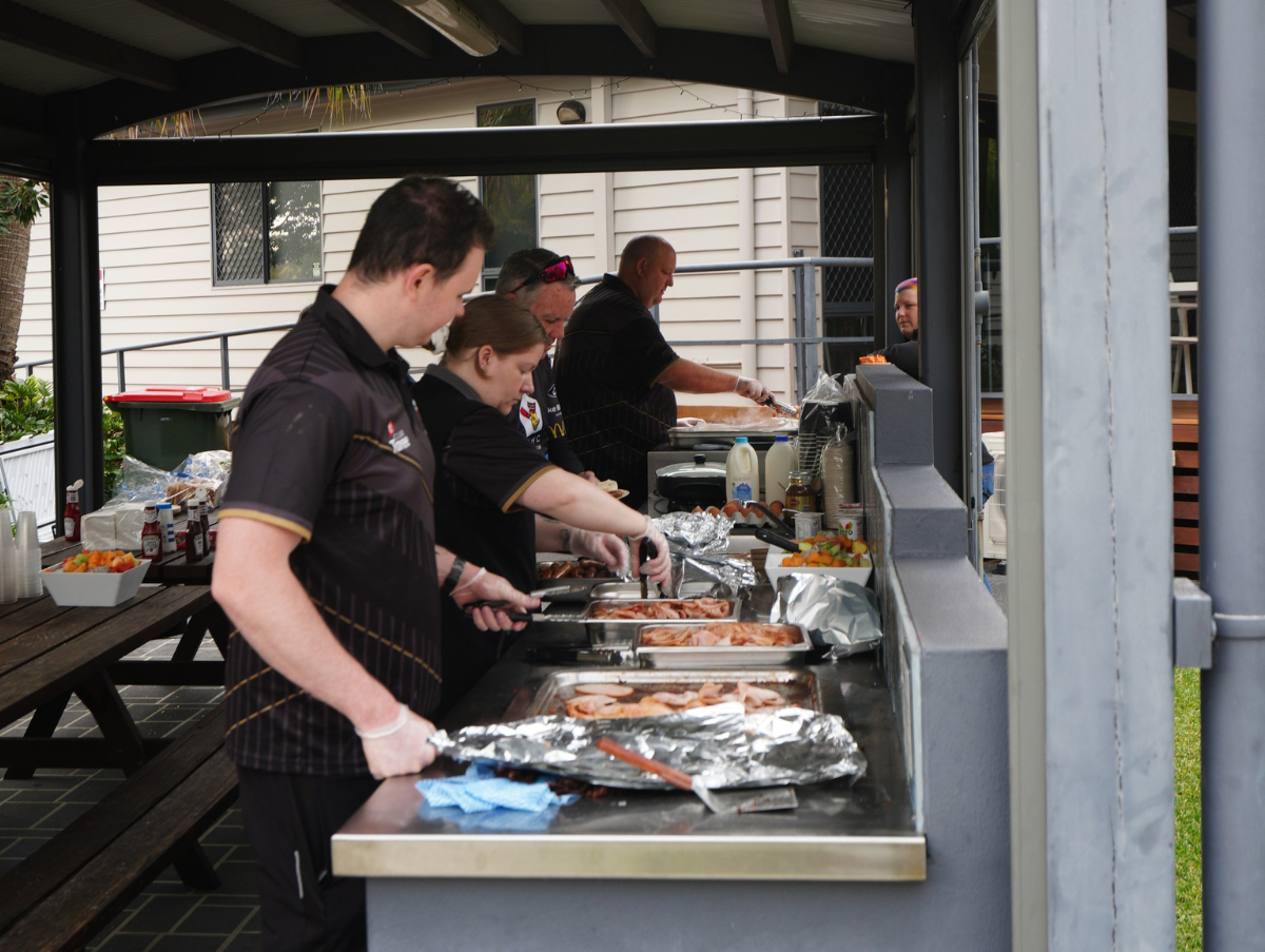 Four adults, all wearing black shirts, cooking a variety of foods on a barbeque at the Forster Family Retreat