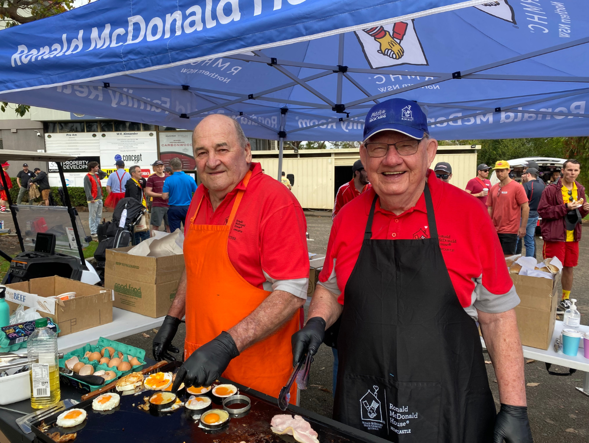 Two elderly gentlemen smile at the camera while standing in front of a barbeque cooking eggs