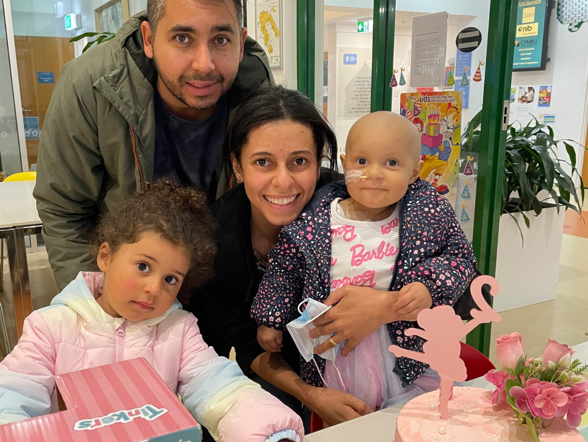 A family (Mum, dad and two young girls) are posing for a photo at Ronald McDonald House Newcastle. The youngest daughter (toddler) is wearing a nasal tube. On a table in front of the family is a pink birthday cake.