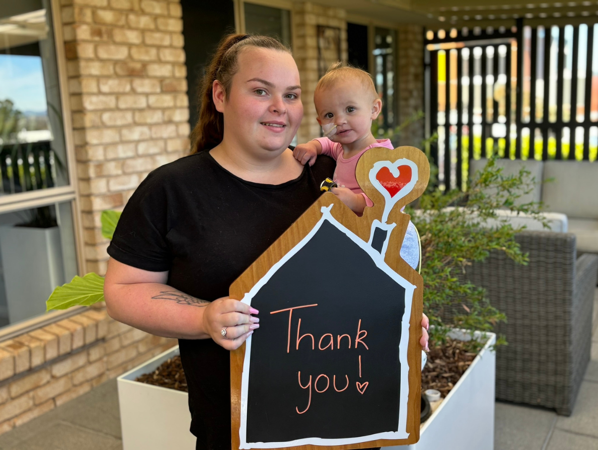 A mum is holding her small daughter outside Ronald McDonald House Tamworth. Her daughter is wearing a nasal tube. In her other hand, the mum is holding a chalk board sign that reads "Thank you!"