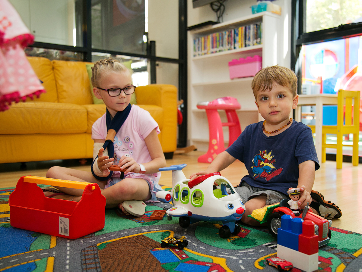 Two kids is sitting in the Ronald McDonald J1 Family Room. Both are playing with a toy plane and tractor. The young girl is wearing black framed glasses and has her right arm in a sling and cast.